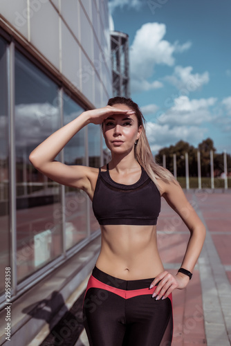 athlete girl in summer in city looks into distance, covering her hand from bright sun. Workout before morning jogging, fitness outdoor workout. Tanned slim figure, motivation woman lifestyle.