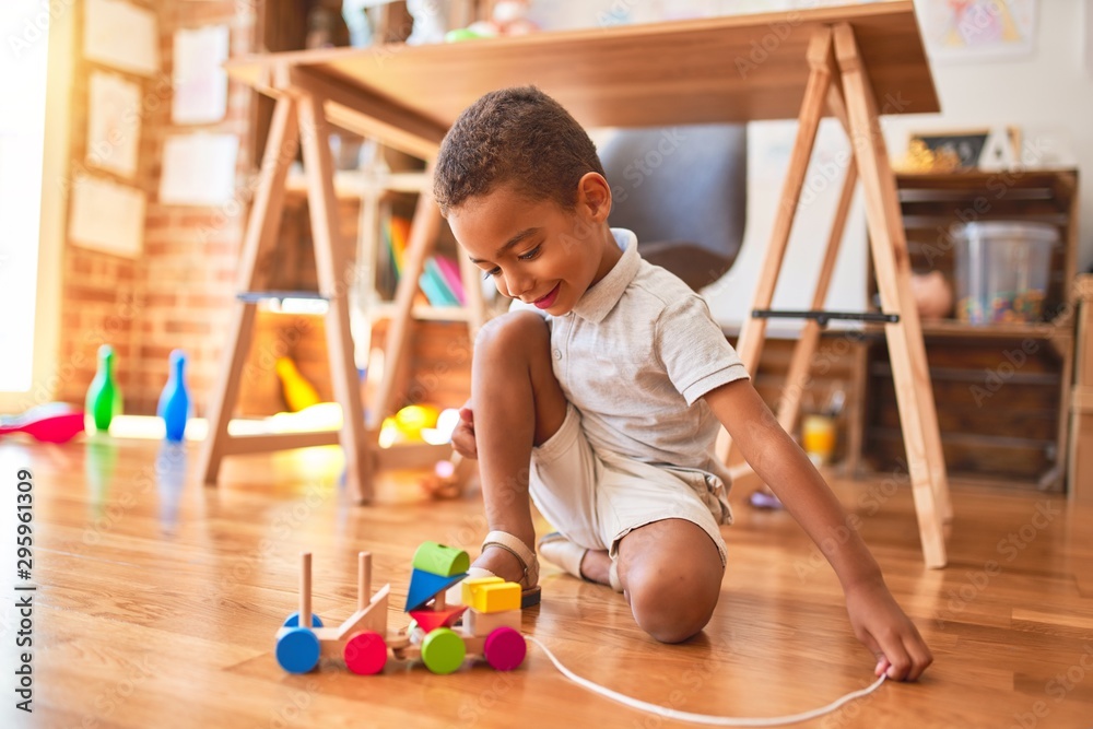 Fototapeta premium Beautiful african american toddler playing with wooden blocks train toy around lots of toys at kindergarten