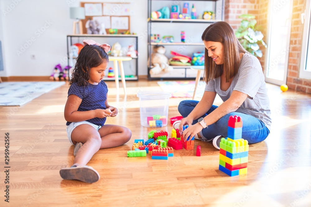 Fototapeta premium Beautiful teacher and toddler girl playing with construction blocks bulding tower at kindergarten