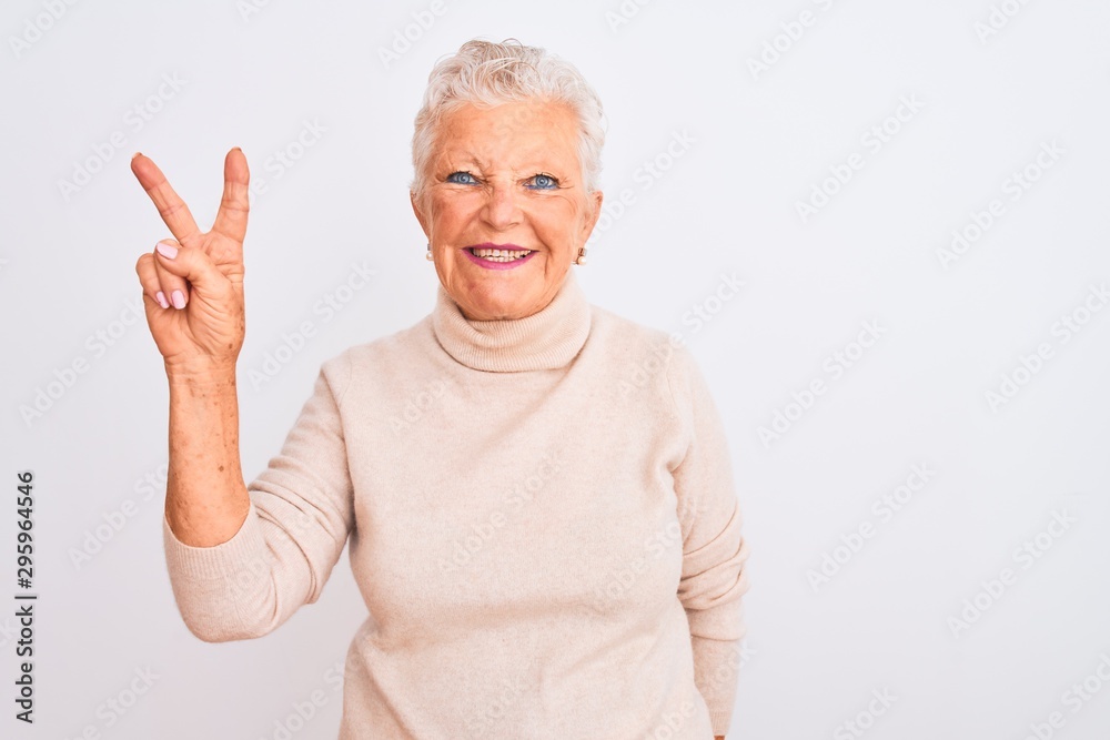 Senior grey-haired woman wearing turtleneck sweater standing over isolated white background smiling with happy face winking at the camera doing victory sign with fingers. Number two. Senior grey-haired woman wearing turtleneck sweater standing over isolated white background smiling with happy face winking at the camera doing victory sign with fingers. Number two.