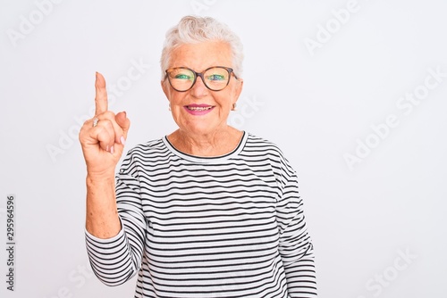 Senior grey-haired woman wearing striped navy t-shirt glasses over isolated white background showing and pointing up with finger number one while smiling confident and happy.