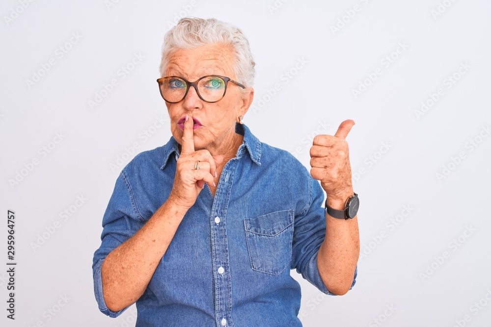 Senior grey-haired woman wearing denim shirt and glasses over isolated white background asking to be quiet with finger on lips pointing with hand to the side. Silence and secret concept. Senior grey-haired woman wearing denim shirt and glasses over isolated white background asking to be quiet with finger on lips pointing with hand to the side. Silence and secret concept.