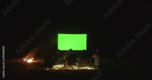 Outdoor cinema. Friends gather in a campfire camp and watch a movie with a projector on the beach near the sea. Green screen. A place for your image.
