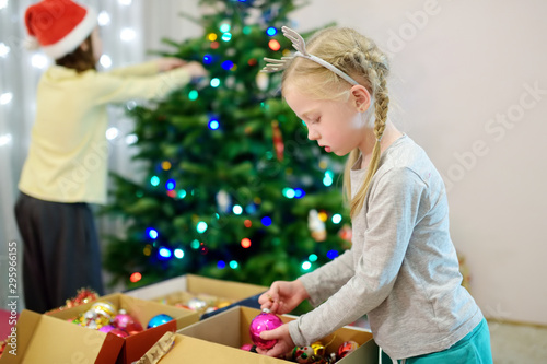 Two adorable sisters decorating a Christmas tree with colorful glass baubles at home. Family leisure at wonderful Xmas time.