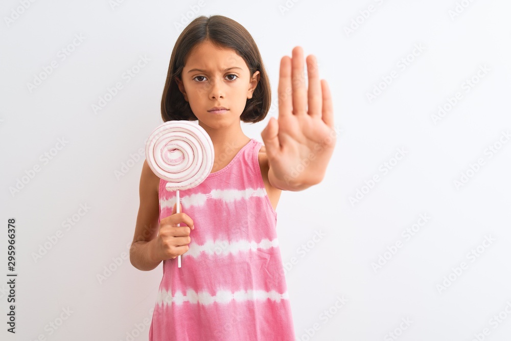 Beautiful child girl eating sweet lollipop standing over isolated white background with open hand doing stop sign with serious and confident expression, defense gesture Beautiful child girl eating sweet lollipop standing over isolated white background with open hand doing stop sign with serious and confident expression, defense gesture