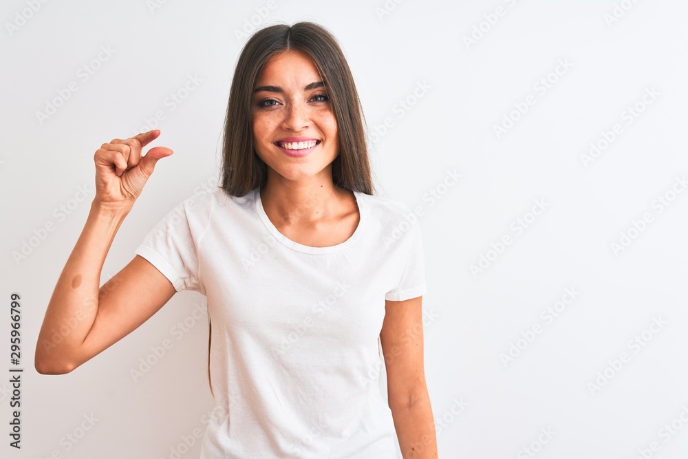 Young beautiful woman wearing casual t-shirt standing over isolated white background smiling and confident gesturing with hand doing small size sign with fingers looking and the camera Young beautiful woman wearing casual t-shirt standing over isolated white background smiling and confident gesturing with hand doing small size sign with fingers looking and the camera