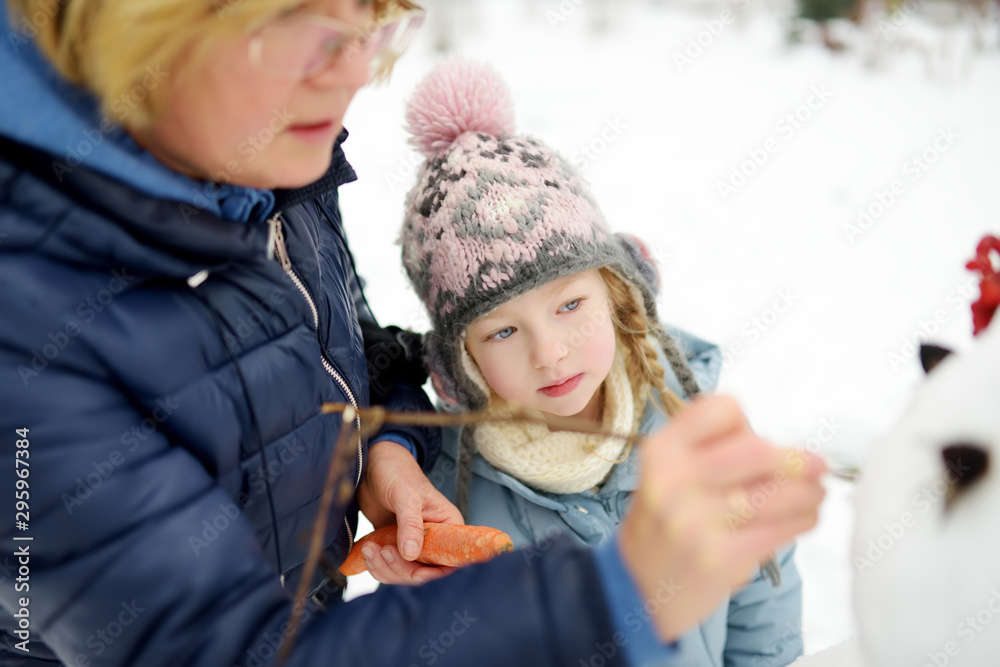 Fototapeta premium Cute little girl and her grandma building a snowman in the backyard. Cute child playing in a snow.