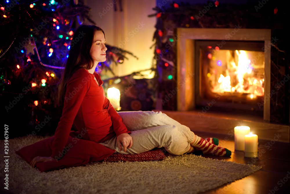 Happy young woman having a good time sitting by a fireplace in a cozy dark living room on Christmas eve. Celebrating Xmas at home.