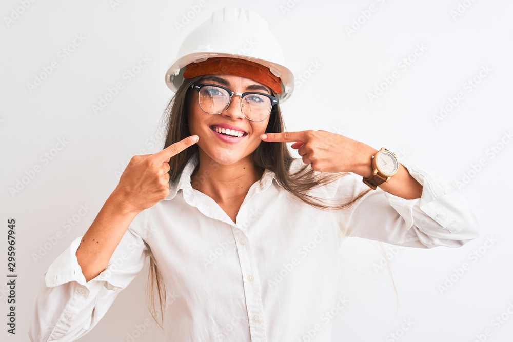 Young beautiful architect woman wearing helmet and glasses over isolated white background smiling cheerful showing and pointing with fingers teeth and mouth. Dental health concept.