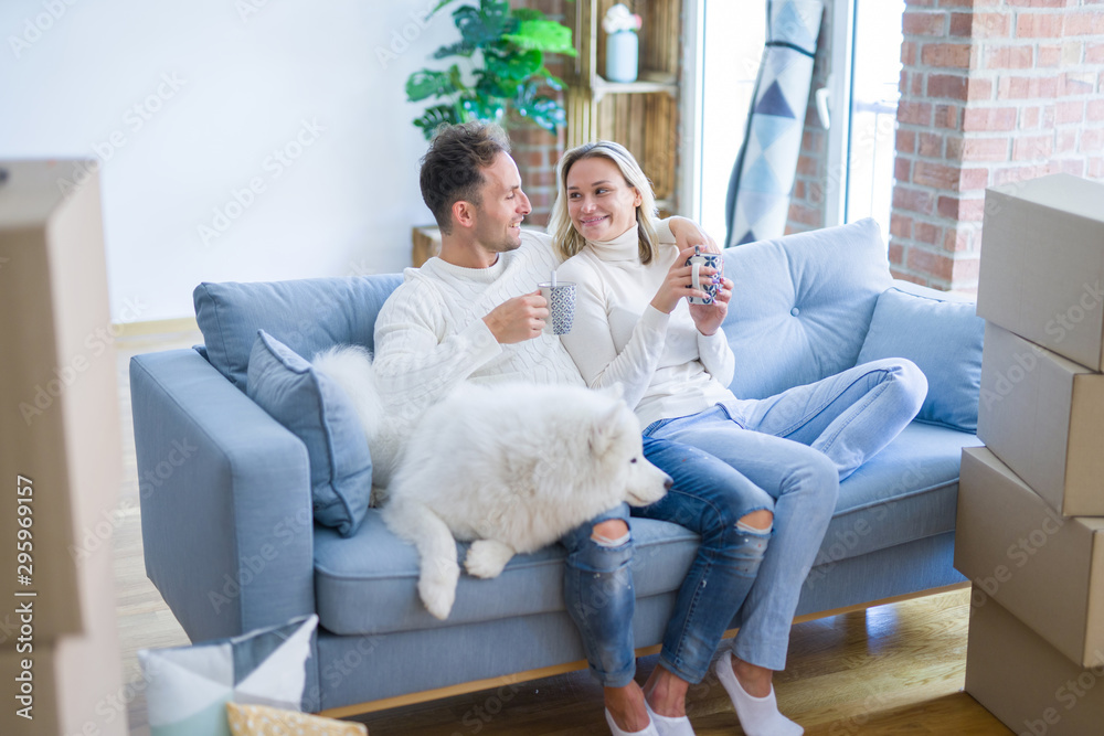 Young beautiful couple with dog sitting on the sofa drinking coffee at new home around cardboard boxes Young beautiful couple with dog sitting on the sofa drinking coffee at new home around cardboard boxes