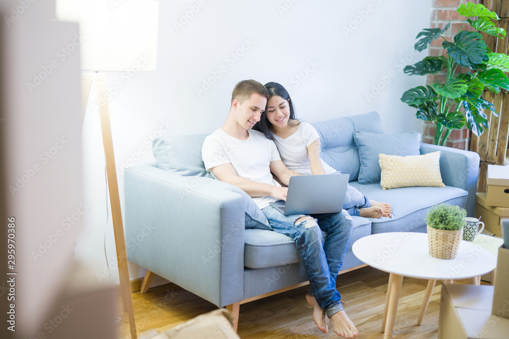 Young beautiful couple sitting on the sofa using laptop at new home around cardboard boxes Young beautiful couple sitting on the sofa using laptop at new home around cardboard boxes