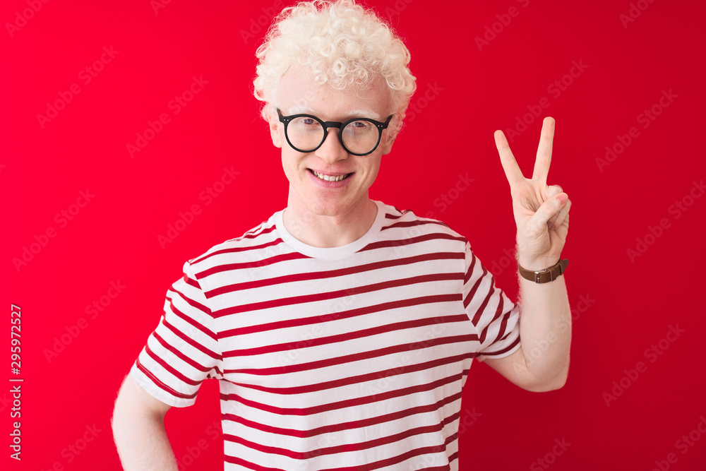 Young albino blond man wearing striped t-shirt and glasses over isolated red background showing and pointing up with fingers number two while smiling confident and happy. Young albino blond man wearing striped t-shirt and glasses over isolated red background showing and pointing up with fingers number two while smiling confident and happy.