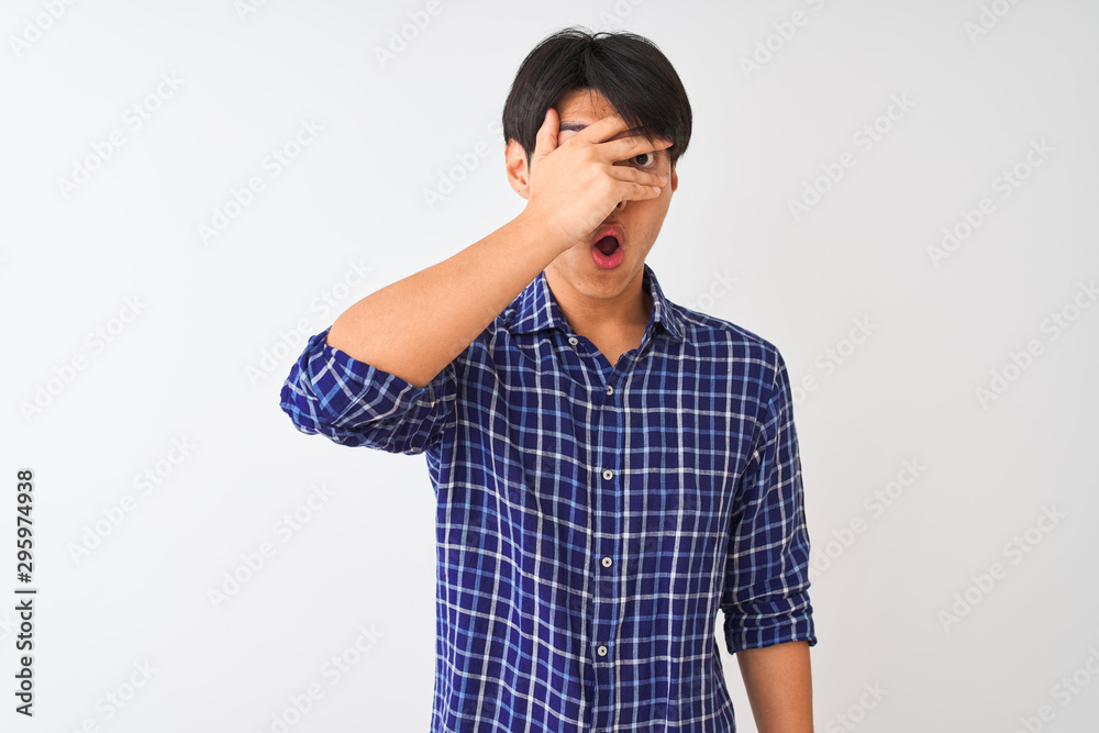 Young chinese man wearing casual blue shirt standing over isolated white background peeking in shock covering face and eyes with hand, looking through fingers with embarrassed expression. Young chinese man wearing casual blue shirt standing over isolated white background peeking in shock covering face and eyes with hand, looking through fingers with embarrassed expression.