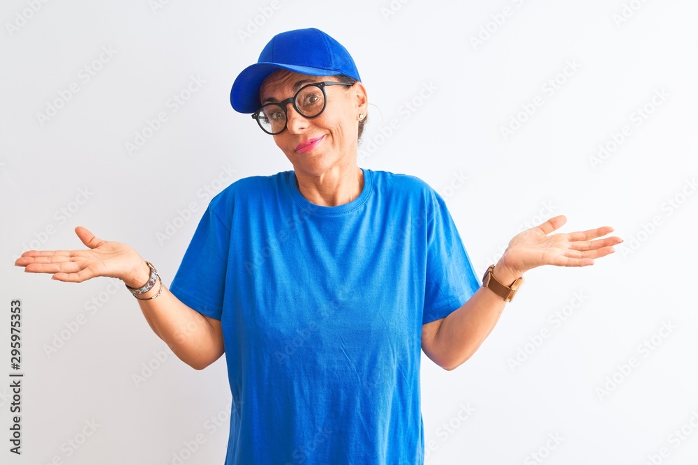 Senior deliverywoman wearing cap and glasses standing over isolated white background clueless and confused expression with arms and hands raised. Doubt concept.