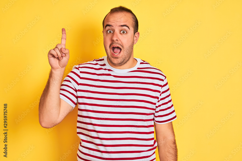 Naklejka premium Young man wearing casual striped t-shirt standing over isolated yellow background pointing finger up with successful idea. Exited and happy. Number one.