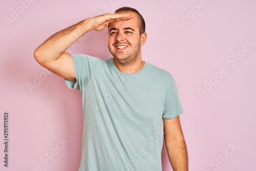 Young man wearing blue casual t-shirt standing over isolated pink background very happy and smiling looking far away with hand over head. Searching concept.