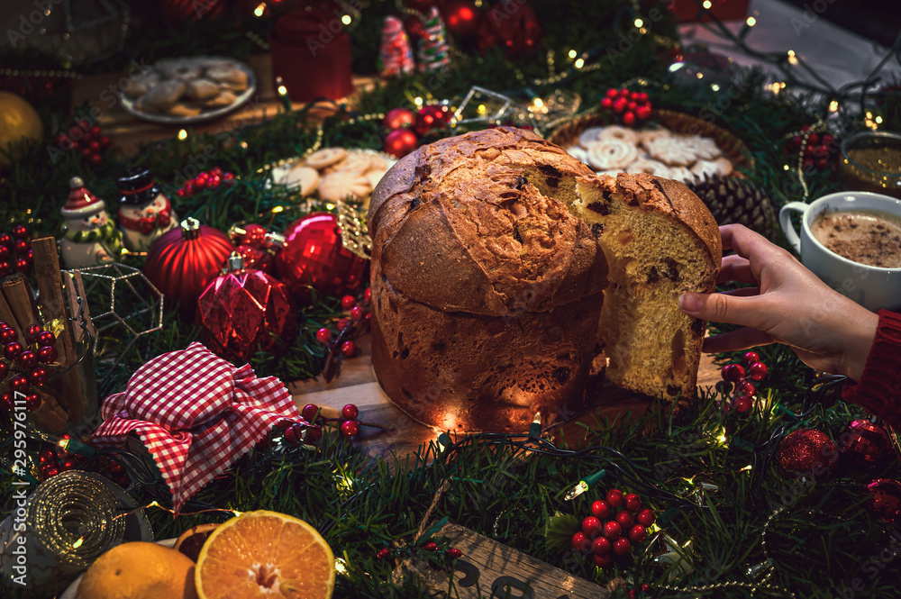 Young woman holding a slice of panettone at Christmas and in the background a table with holiday season ingredients like hot chocolate, orange, cookies, cinnamon and pine