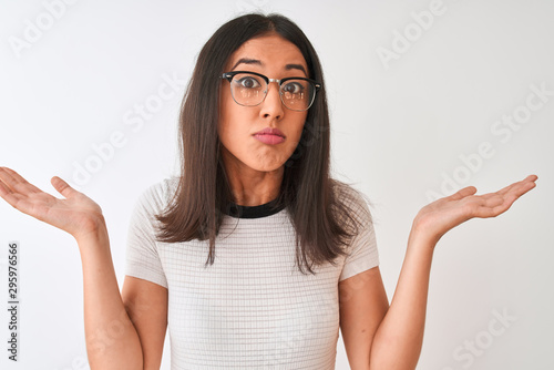Chinese woman wearing casual t-shirt and glasses standing over isolated white background clueless and confused expression with arms and hands raised. Doubt concept.