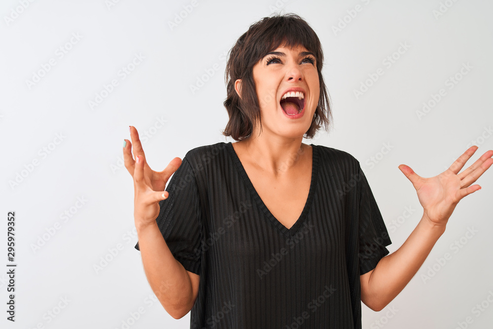 Young beautiful woman wearing black t-shirt standing over isolated white background crazy and mad shouting and yelling with aggressive expression and arms raised. Frustration concept. Young beautiful woman wearing black t-shirt standing over isolated white background crazy and mad shouting and yelling with aggressive expression and arms raised. Frustration concept.