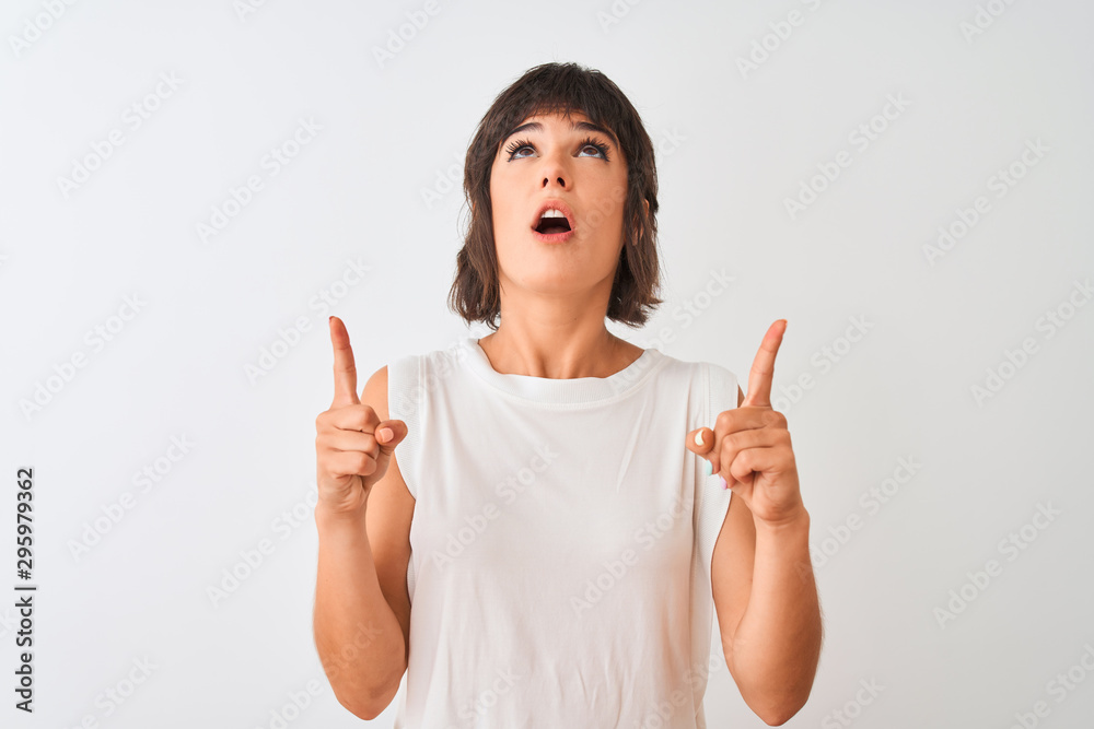 Young beautiful woman wearing casual t-shirt standing over isolated white background amazed and surprised looking up and pointing with fingers and raised arms. Young beautiful woman wearing casual t-shirt standing over isolated white background amazed and surprised looking up and pointing with fingers and raised arms.