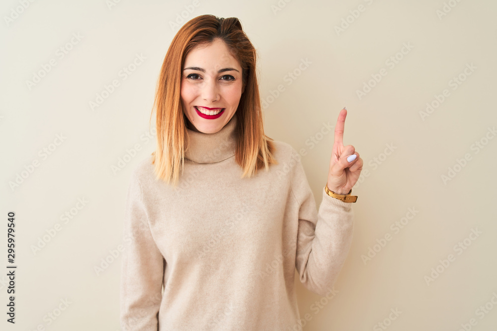 Redhead woman wearing elegant turtleneck sweater standing over isolated white background showing and pointing up with finger number one while smiling confident and happy. Redhead woman wearing elegant turtleneck sweater standing over isolated white background showing and pointing up with finger number one while smiling confident and happy.
