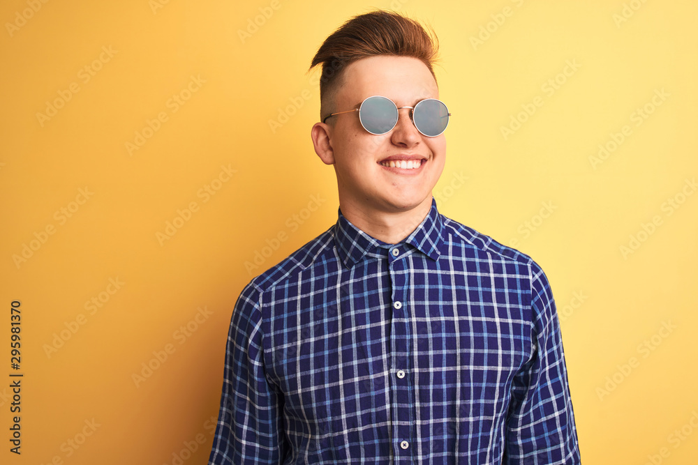 Young handsome man wearing casual shirt and sunglasses over isolated yellow background looking away to side with smile on face, natural expression. Laughing confident. Young handsome man wearing casual shirt and sunglasses over isolated yellow background looking away to side with smile on face, natural expression. Laughing confident.