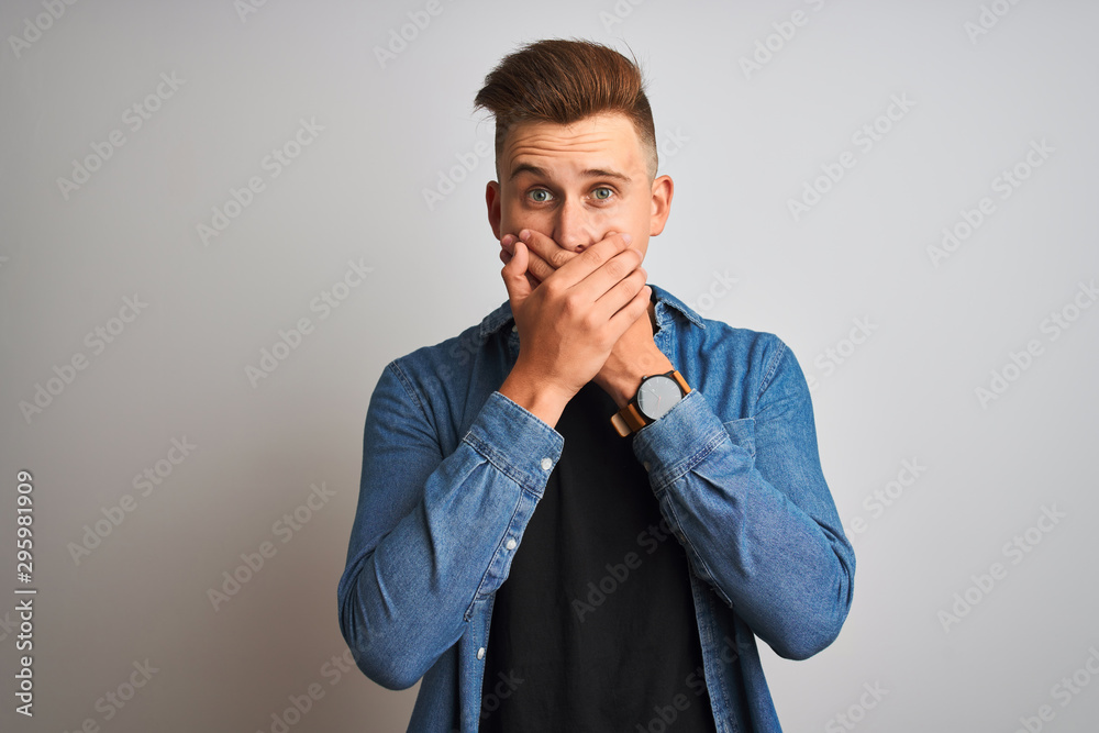 Young handsome man wearing denim shirt standing over isolated white background shocked covering mouth with hands for mistake. Secret concept.