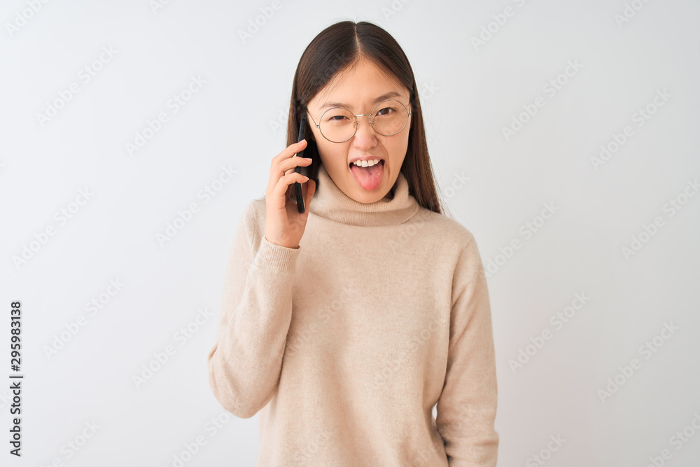 Young chinese woman talking on the smartphone over isolated white background sticking tongue out happy with funny expression. Emotion concept. Young chinese woman talking on the smartphone over isolated white background sticking tongue out happy with funny expression. Emotion concept.