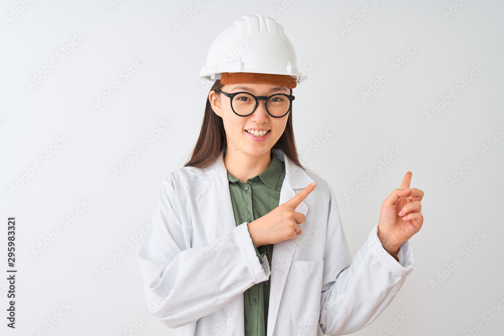 Young chinese engineer woman wearing coat helmet glasses over isolated white background smiling and looking at the camera pointing with two hands and fingers to the side. Young chinese engineer woman wearing coat helmet glasses over isolated white background smiling and looking at the camera pointing with two hands and fingers to the side.