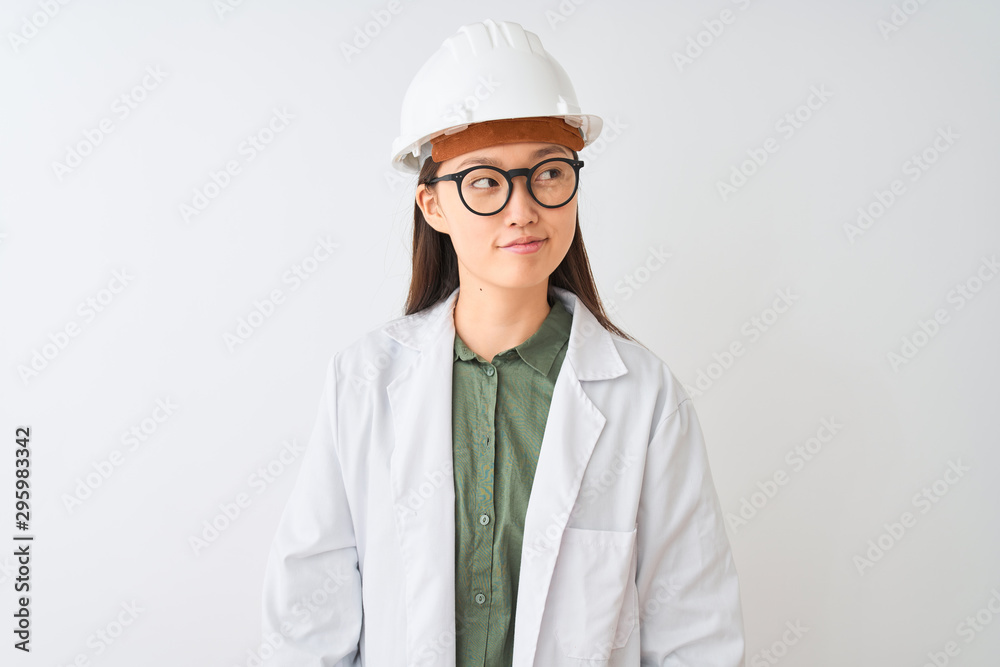 Young chinese engineer woman wearing coat helmet glasses over isolated white background smiling looking to the side and staring away thinking. Young chinese engineer woman wearing coat helmet glasses over isolated white background smiling looking to the side and staring away thinking.