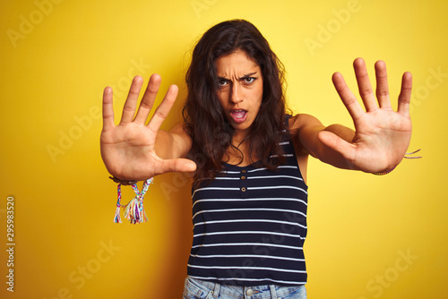 Young beautiful woman wearing striped t-shirt standing over isolated yellow background doing stop gesture with hands palms, angry and frustration expression