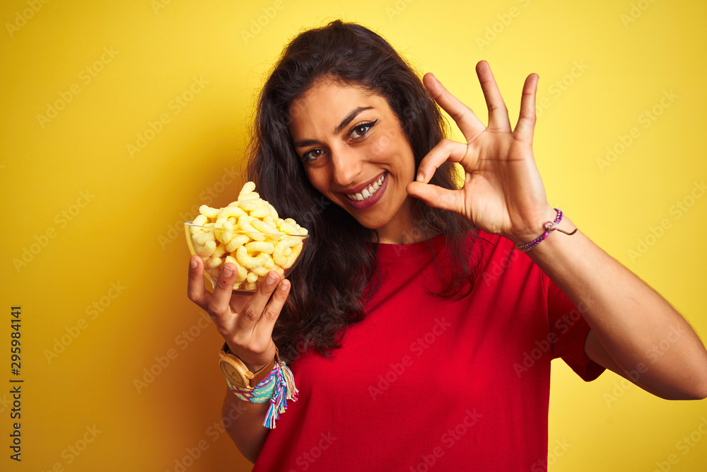 Young beautiful woman holding bowl with extruded corn over isolated yellow background doing ok sign with fingers, excellent symbol Young beautiful woman holding bowl with extruded corn over isolated yellow background doing ok sign with fingers, excellent symbol