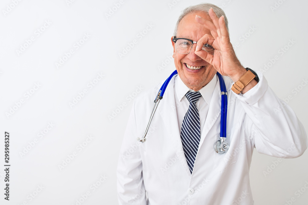 Senior grey-haired doctor man wearing stethoscope standing over isolated white background doing ok gesture with hand smiling, eye looking through fingers with happy face. Senior grey-haired doctor man wearing stethoscope standing over isolated white background doing ok gesture with hand smiling, eye looking through fingers with happy face.