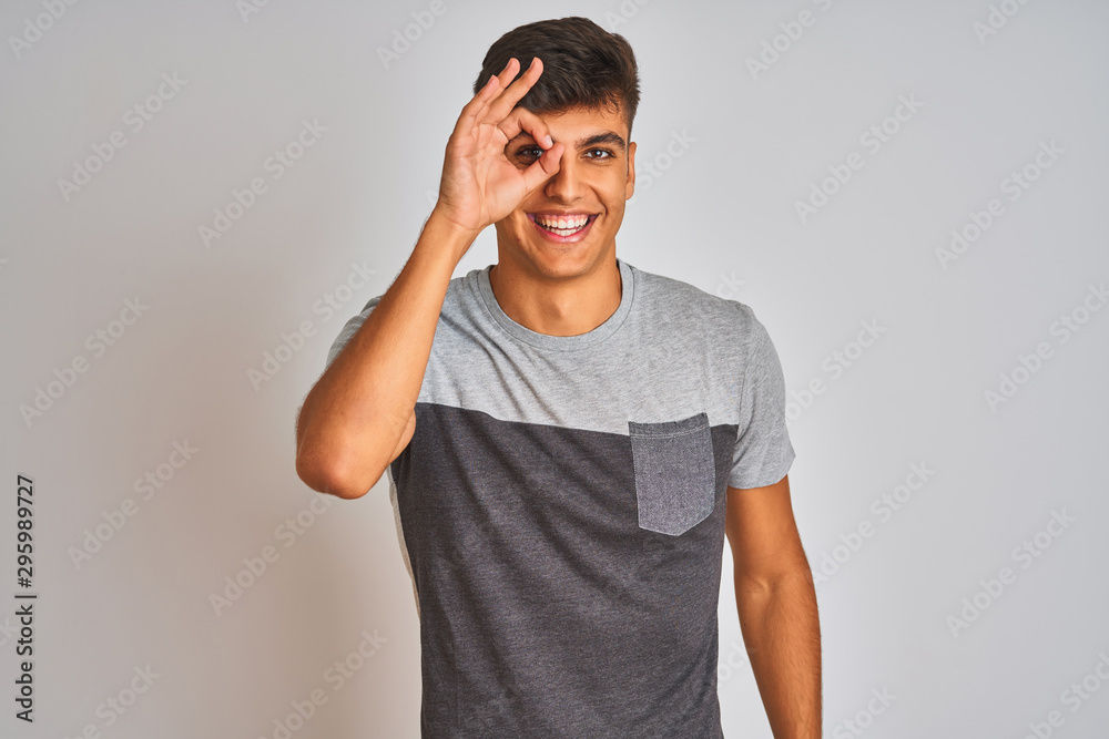 Young indian man wearing casual t-shirt standing over isolated white background doing ok gesture with hand smiling, eye looking through fingers with happy face. Young indian man wearing casual t-shirt standing over isolated white background doing ok gesture with hand smiling, eye looking through fingers with happy face.