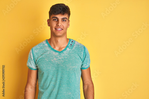 Young indian man wearing green t-shirt standing over isolated yellow background with a happy and cool smile on face. Lucky person.