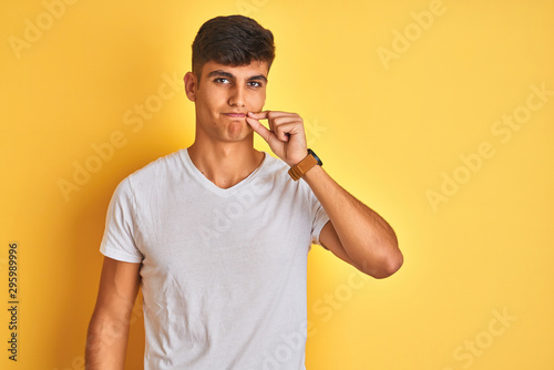 Young indian man wearing white t-shirt standing over isolated yellow background mouth and lips shut as zip with fingers. Secret and silent  taboo talking