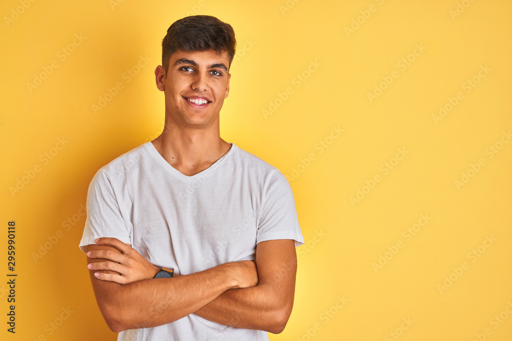 Young indian man wearing white t-shirt standing over isolated yellow background happy face smiling with crossed arms looking at the camera. Positive person. Young indian man wearing white t-shirt standing over isolated yellow background happy face smiling with crossed arms looking at the camera. Positive person.