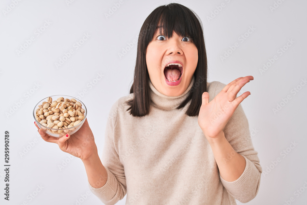 Young beautiful Chinese woman holding bowl with pistachios over isolated white background very happy and excited, winner expression celebrating victory screaming with big smile and raised hands Young beautiful Chinese woman holding bowl with pistachios over isolated white background very happy and excited, winner expression celebrating victory screaming with big smile and raised hands