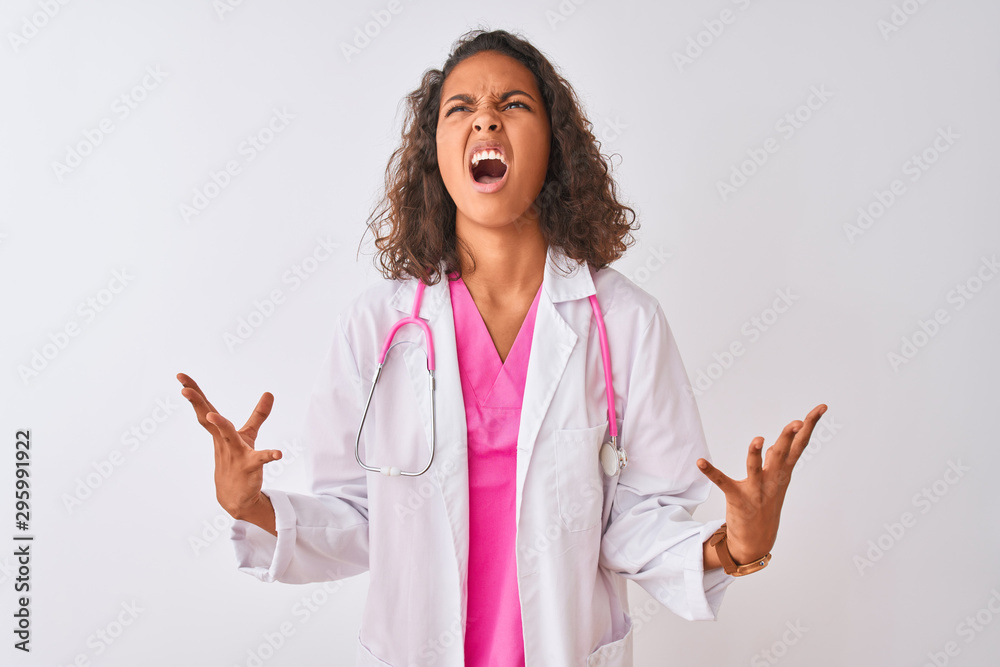 Young brazilian doctor woman wearing stethoscope standing over isolated white background crazy and mad shouting and yelling with aggressive expression and arms raised. Frustration concept. Young brazilian doctor woman wearing stethoscope standing over isolated white background crazy and mad shouting and yelling with aggressive expression and arms raised. Frustration concept.