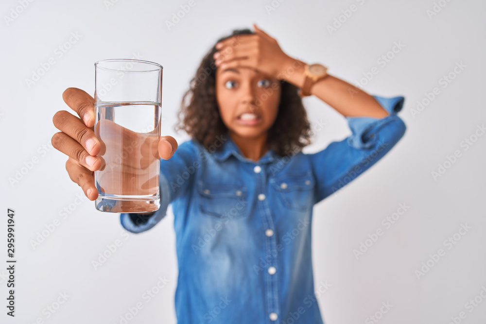 Young brazilian woman holding glass of water standing over isolated white background stressed with hand on head, shocked with shame and surprise face, angry and frustrated. Fear and upset for mistake. Young brazilian woman holding glass of water standing over isolated white background stressed with hand on head, shocked with shame and surprise face, angry and frustrated. Fear and upset for mistake.