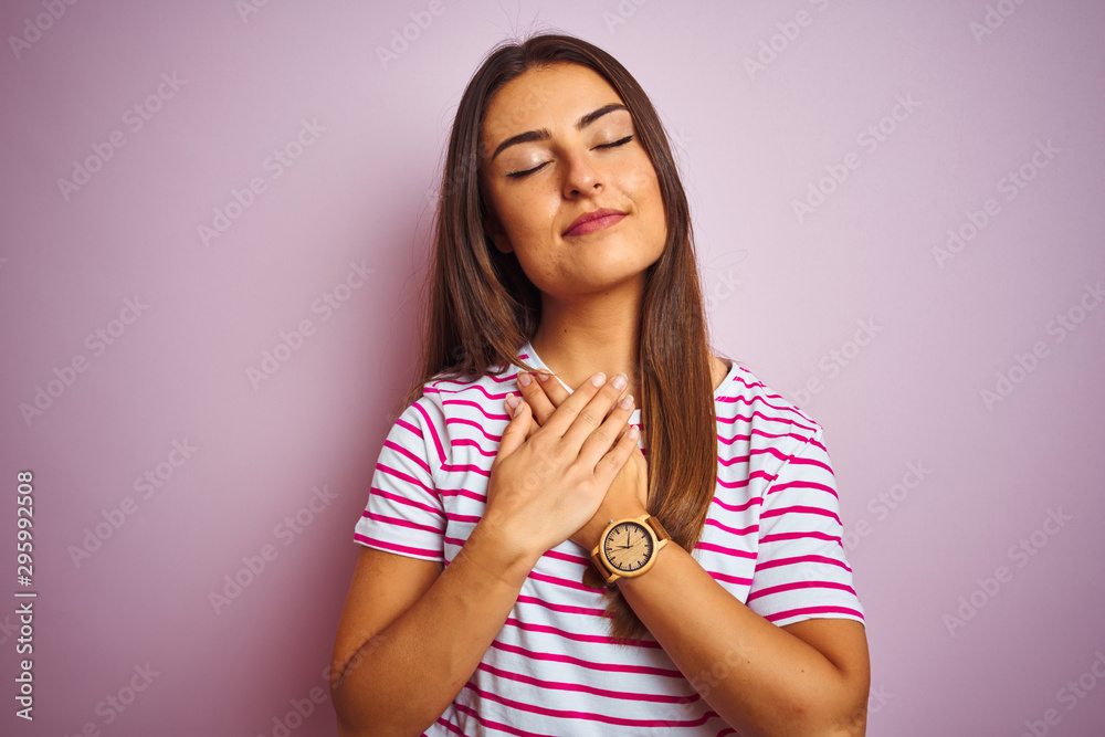 Young beautiful woman wearing striped t-shirt standing over isolated pink background smiling with hands on chest with closed eyes and grateful gesture on face. Health concept. Young beautiful woman wearing striped t-shirt standing over isolated pink background smiling with hands on chest with closed eyes and grateful gesture on face. Health concept.