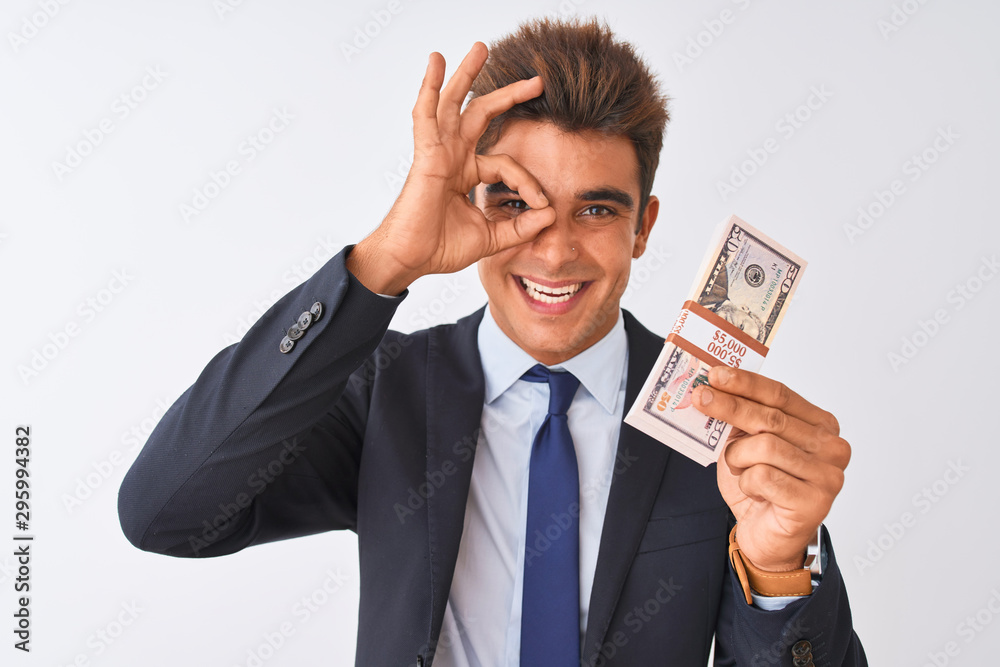 Young handsome businessman wearing suit holding dollars over isolated white background with happy face smiling doing ok sign with hand on eye looking through fingers Young handsome businessman wearing suit holding dollars over isolated white background with happy face smiling doing ok sign with hand on eye looking through fingers