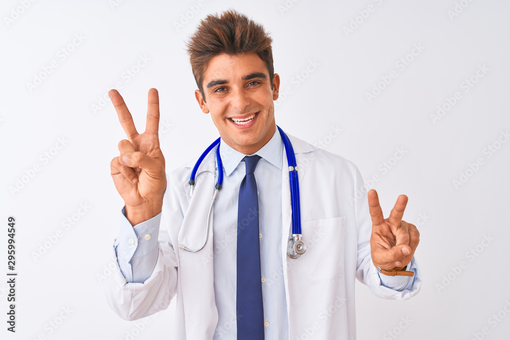 Young handsome doctor man wearing stethoscope over isolated white background smiling looking to the camera showing fingers doing victory sign. Number two. Young handsome doctor man wearing stethoscope over isolated white background smiling looking to the camera showing fingers doing victory sign. Number two.