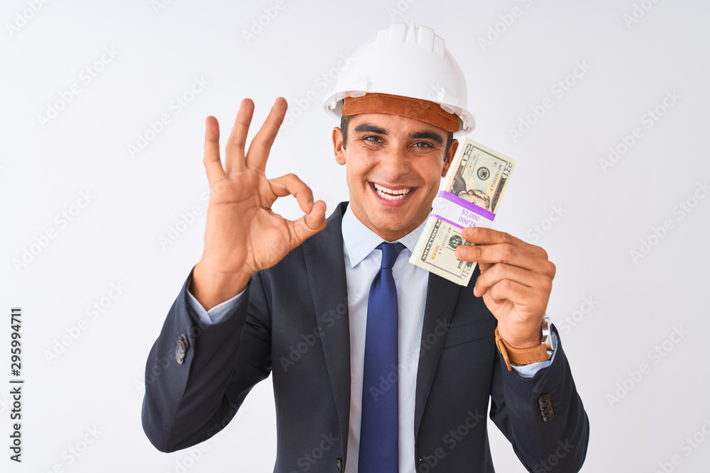 Young handsome architect man wearing helmet holding dollars over isolated white background doing ok sign with fingers, excellent symbol Young handsome architect man wearing helmet holding dollars over isolated white background doing ok sign with fingers, excellent symbol