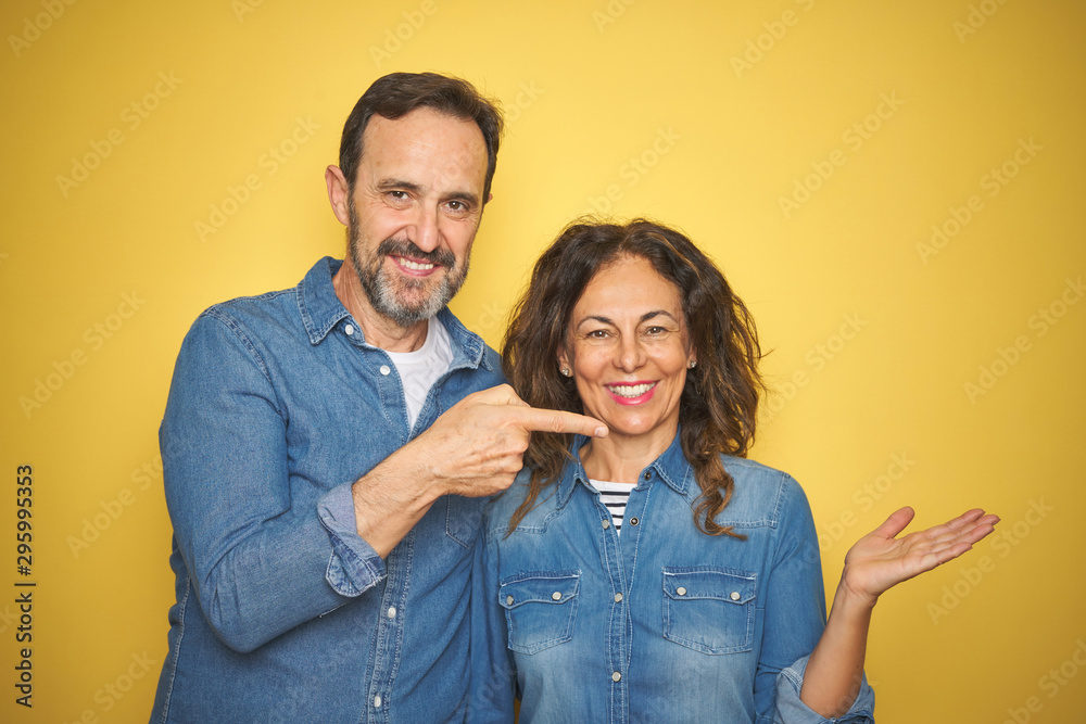 Obraz premium Beautiful middle age couple together standing over isolated yellow background amazed and smiling to the camera while presenting with hand and pointing with finger.