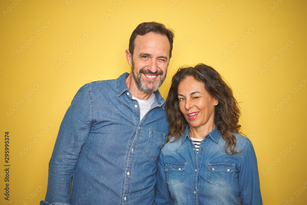 Beautiful middle age couple together standing over isolated yellow background winking looking at the camera with sexy expression, cheerful and happy face. Beautiful middle age couple together standing over isolated yellow background winking looking at the camera with sexy expression, cheerful and happy face.