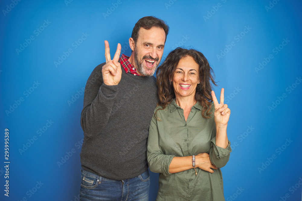 Beautiful middle age couple together standing over isolated blue background smiling with happy face winking at the camera doing victory sign. Number two. Beautiful middle age couple together standing over isolated blue background smiling with happy face winking at the camera doing victory sign. Number two.