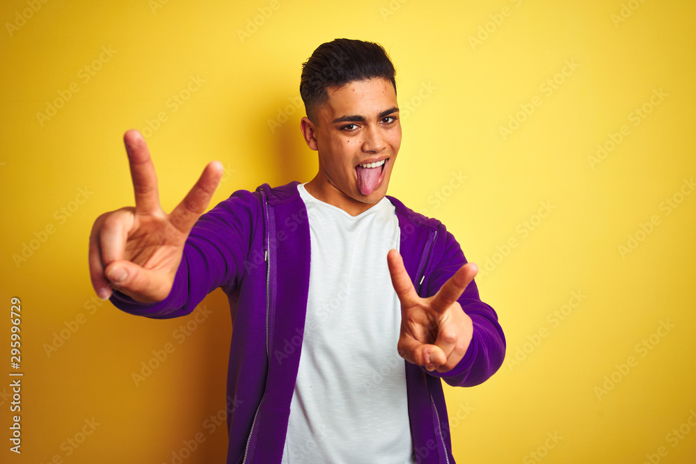 Young brazilian man wearing purple sweatshirt standing over isolated yellow background smiling with tongue out showing fingers of both hands doing victory sign. Number two. Young brazilian man wearing purple sweatshirt standing over isolated yellow background smiling with tongue out showing fingers of both hands doing victory sign. Number two.