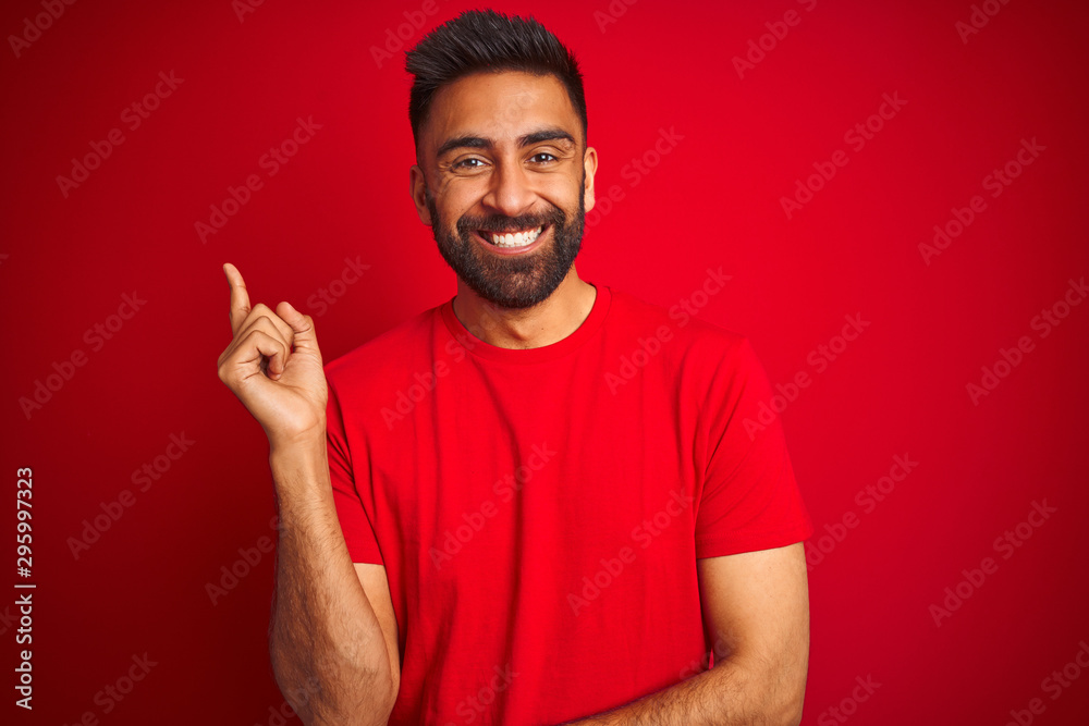 Young handsome indian man wearing t-shirt over isolated red background with a big smile on face, pointing with hand and finger to the side looking at the camera. Young handsome indian man wearing t-shirt over isolated red background with a big smile on face, pointing with hand and finger to the side looking at the camera.
