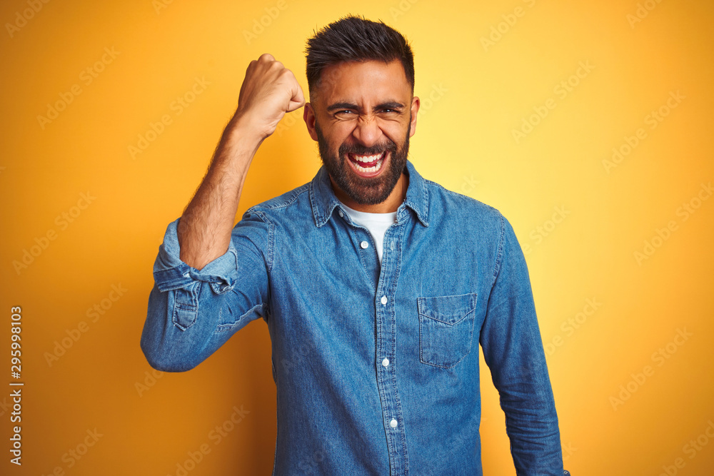 Young indian man wearing denim shirt standing over isolated yellow background angry and mad raising fist frustrated and furious while shouting with anger. Rage and aggressive concept. Young indian man wearing denim shirt standing over isolated yellow background angry and mad raising fist frustrated and furious while shouting with anger. Rage and aggressive concept.